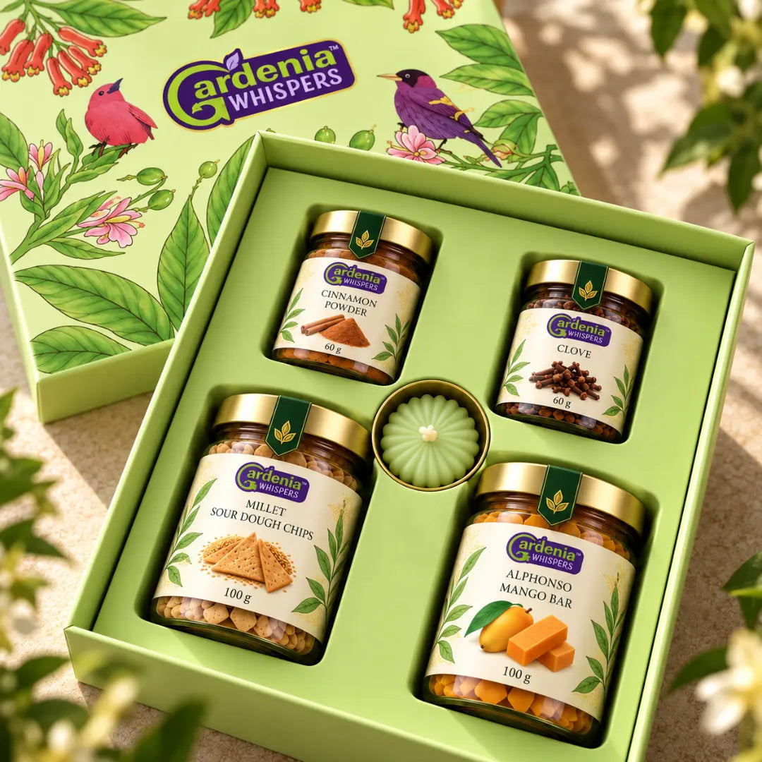 Gardenia Whispers Parijat gift box overhead angle showing four jars of cinnamon powder, clove, millet sour dough chips and alphonso mango bar with green rosette centrepiece, green botanical lid visible above, loose cloves and greenery around