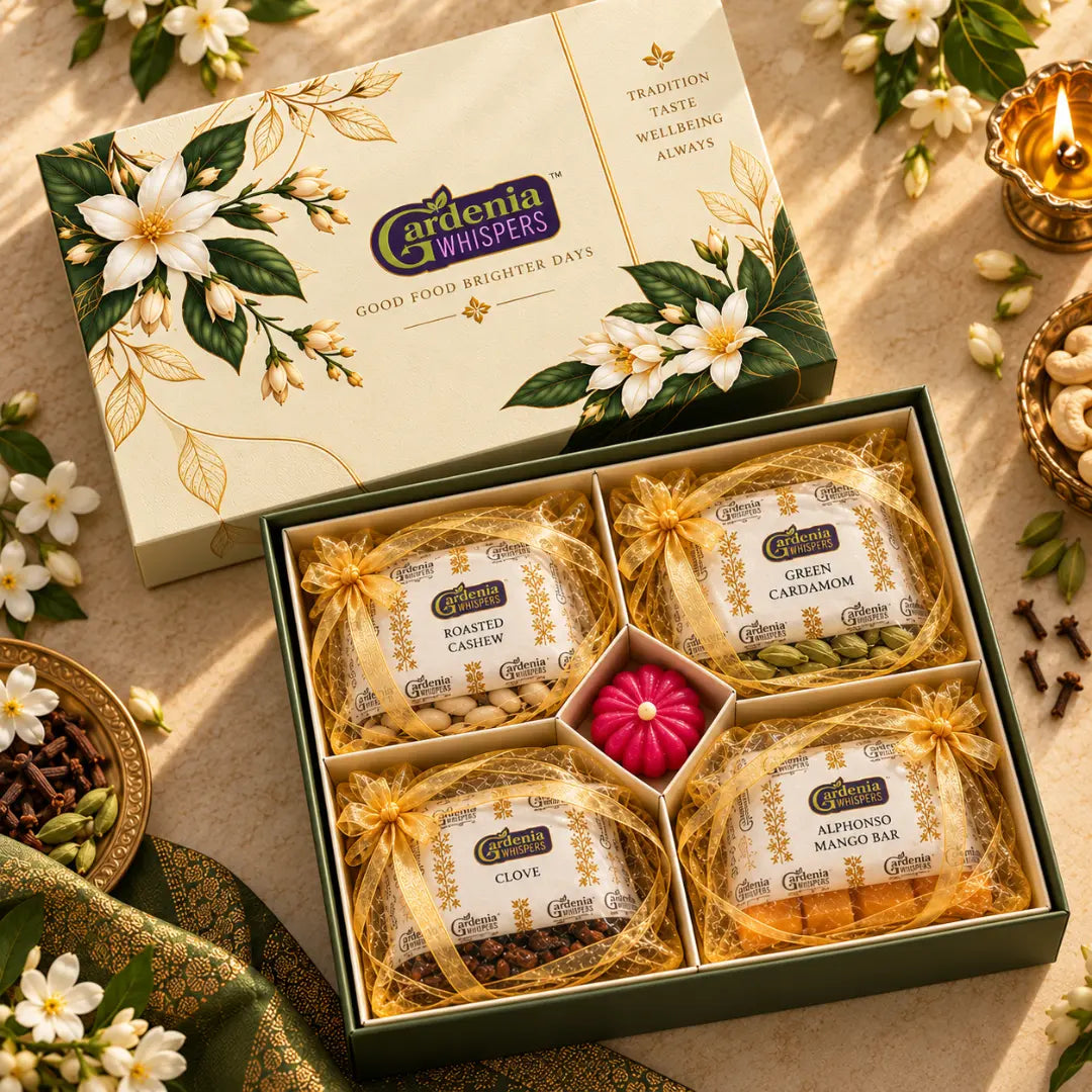 Gardenia Whispers Shephali festive gift box overhead angle with roasted cashew, green cardamom, clove and alphonso mango bar potli pouches arranged around a pink centrepiece, green silk fabric and brass diya in background