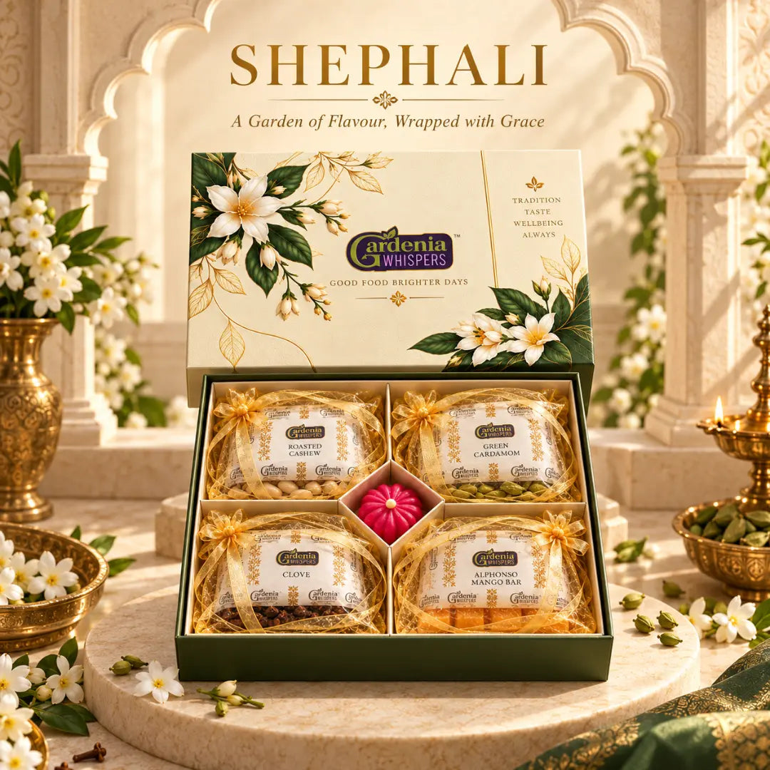Gardenia Whispers Shephali gift box open on marble pedestal showing four gold potli pouches of roasted cashew, green cardamom, clove and alphonso mango bar with a pink decorative centrepiece, floral lid propped behind, set against an Indian arch backdrop with diyas and jasmine